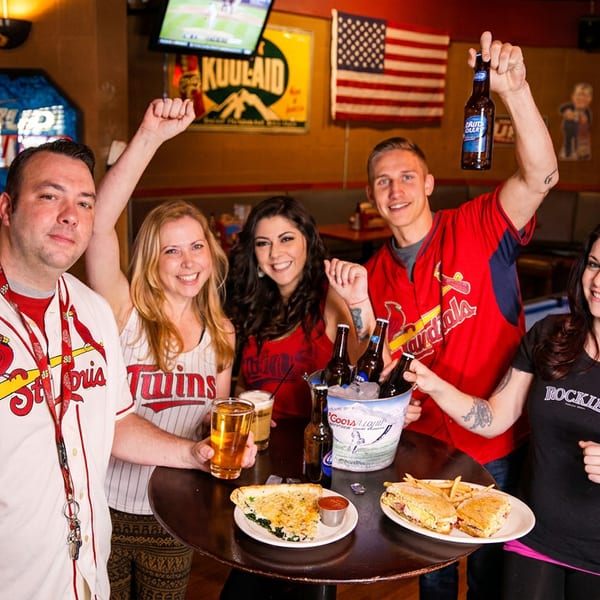 Five smiling friends in sports jerseys celebrating with drinks and food at a bar.