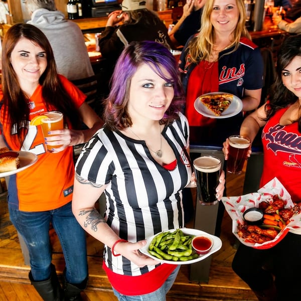 Four women in sports jerseys holding hot dogs, pizza, edamame, and chicken wings in a bar.