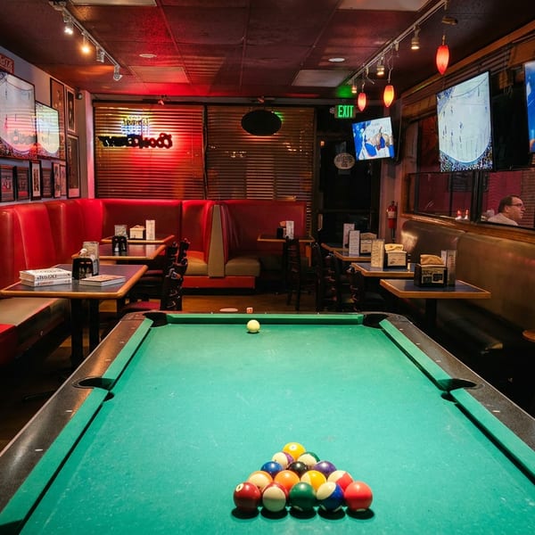 A pool table with racked balls in a dimly lit bar with red booths and TVs showing sports.