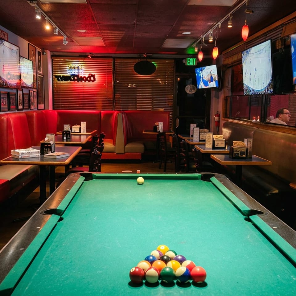 A pool table with racked balls in a dimly lit bar with red booths and TVs showing sports.