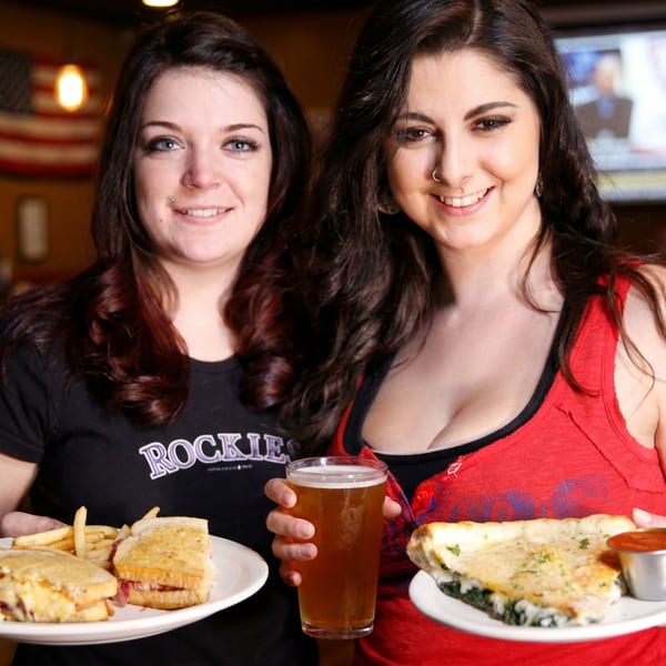 Two smiling women holding plates of sandwiches, fries, a quesadilla, and a beer at a bar.