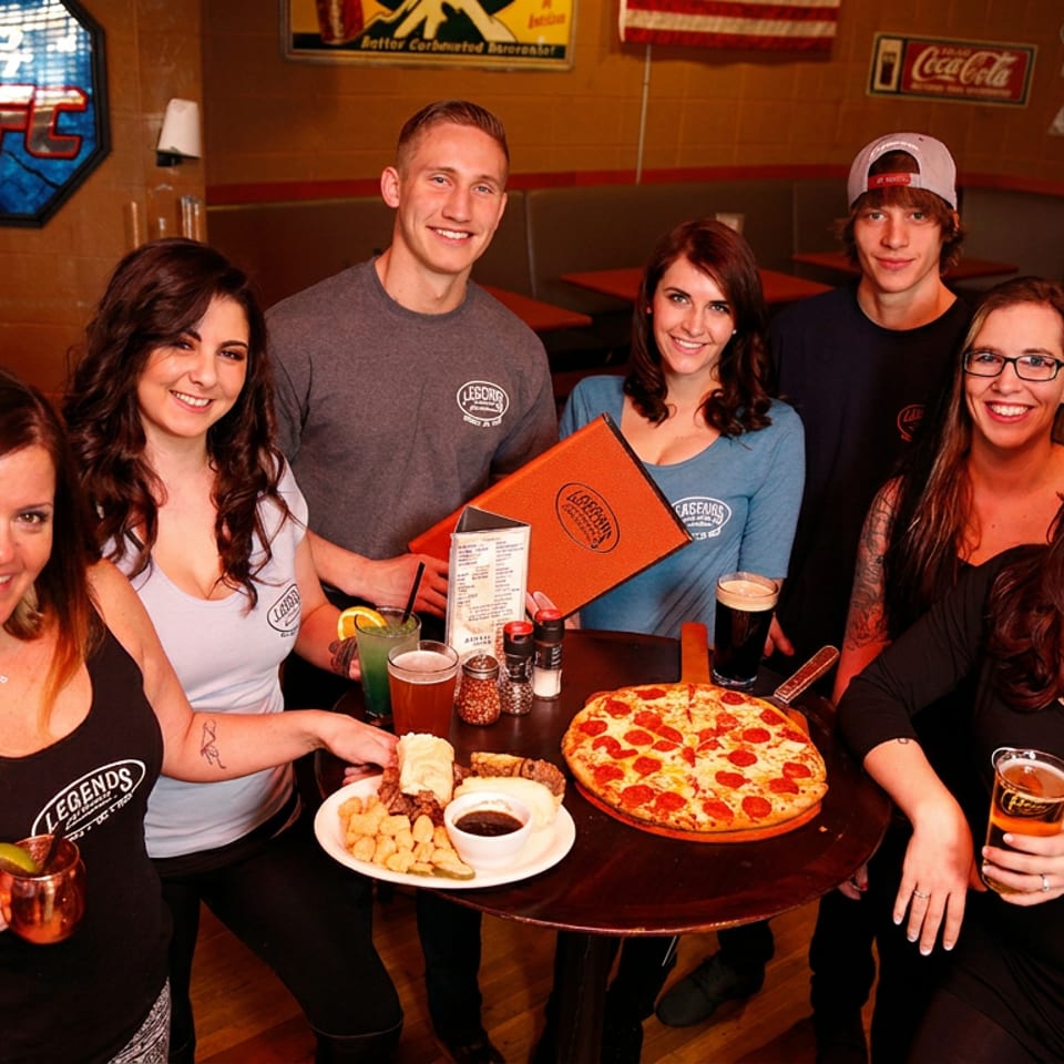 Seven smiling people, including staff and patrons, gathered around a table with pizza, food, and drinks.
