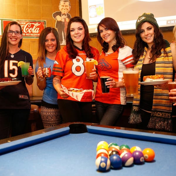 Six women in sports jerseys pose with drinks and food around a pool table in a bar.