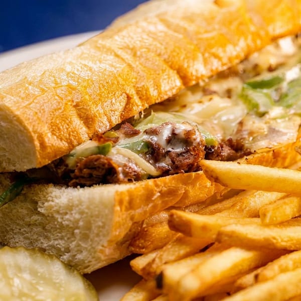 Cheesesteak sandwich with melted cheese, peppers, and onions, served with french fries and pickles.