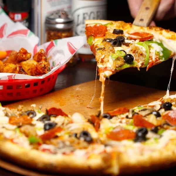 A hand lifts a slice of pepperoni, olive, and pepper pizza with melted cheese, next to a basket of wings.
