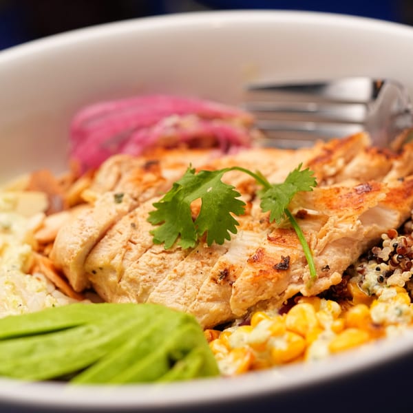 Bowl of sliced grilled chicken with quinoa, corn, rice, greens, and cilantro garnish.