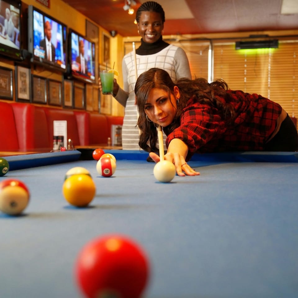 Two women playing pool in a lively bar with red booths and TVs on the wall.