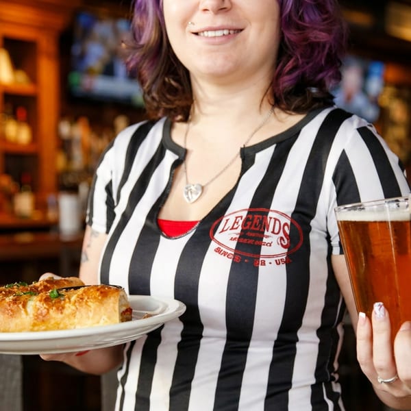 Smiling server in a striped shirt holding a plate of food and two beers at Legends.