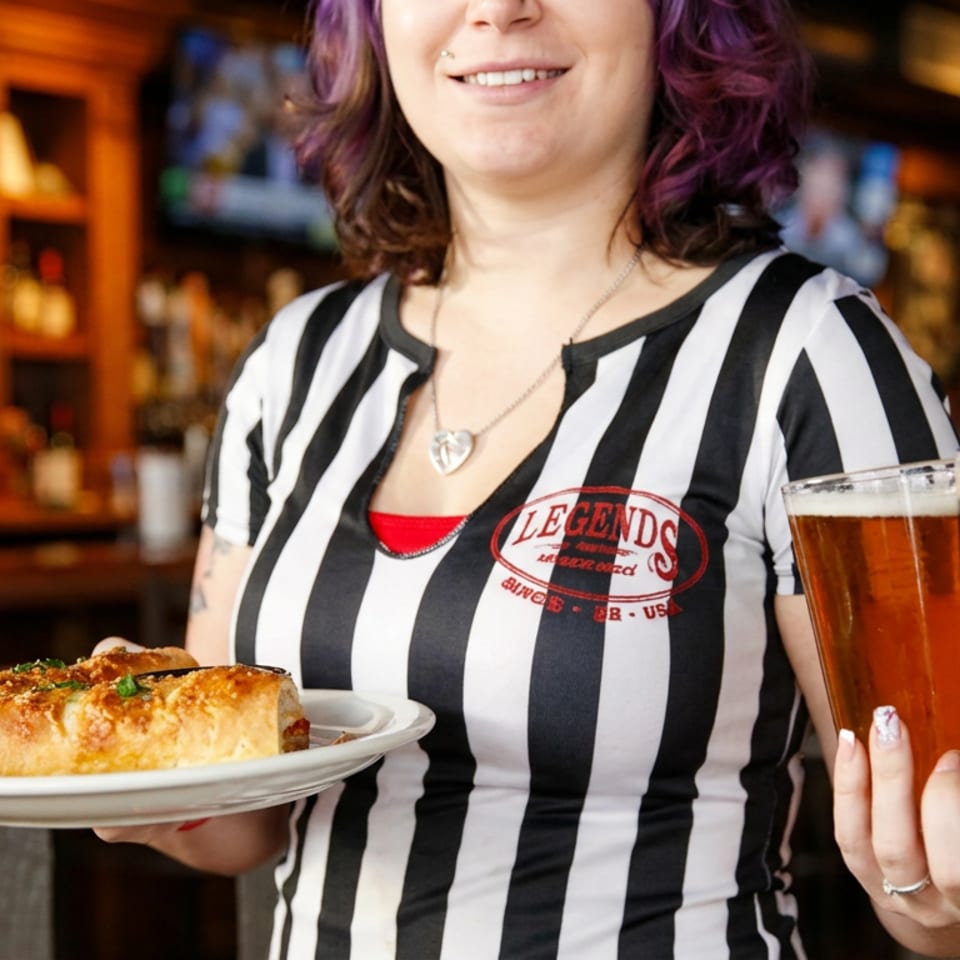 Smiling server in a striped shirt holding a plate of food and two beers at Legends.