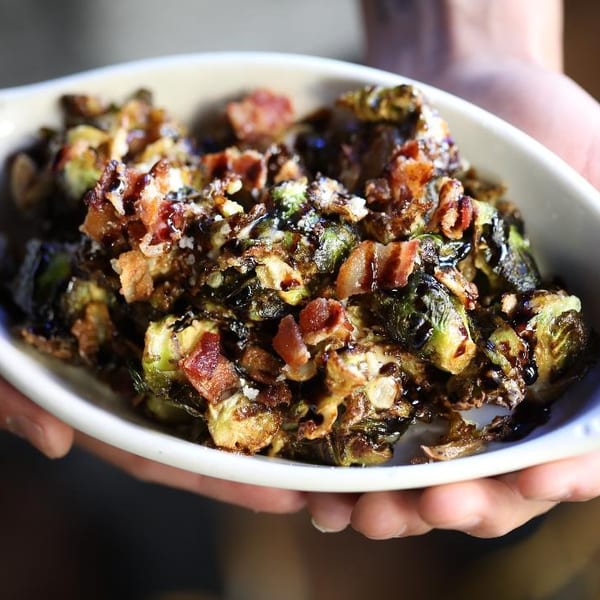 Roasted Brussels sprouts topped with crispy bacon and glaze, served in a white dish held by hands.