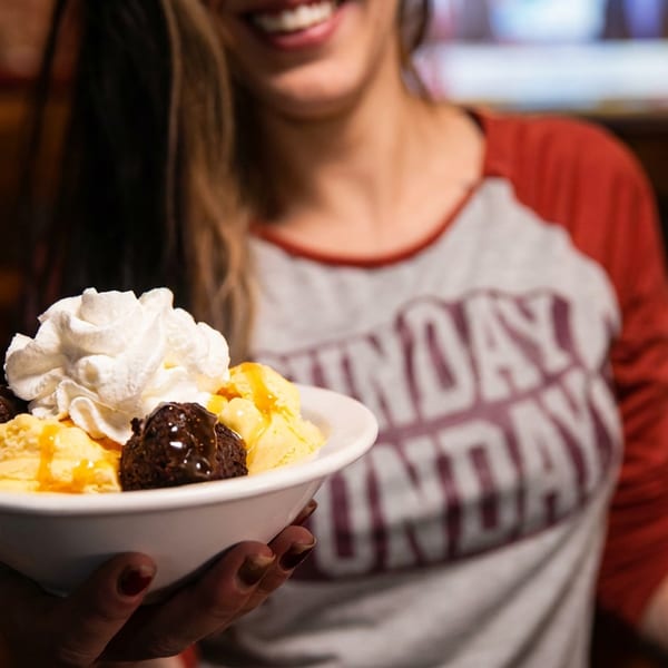 Smiling person holding a bowl of ice cream with whipped cream, chocolate brownie bites, and caramel drizzle.